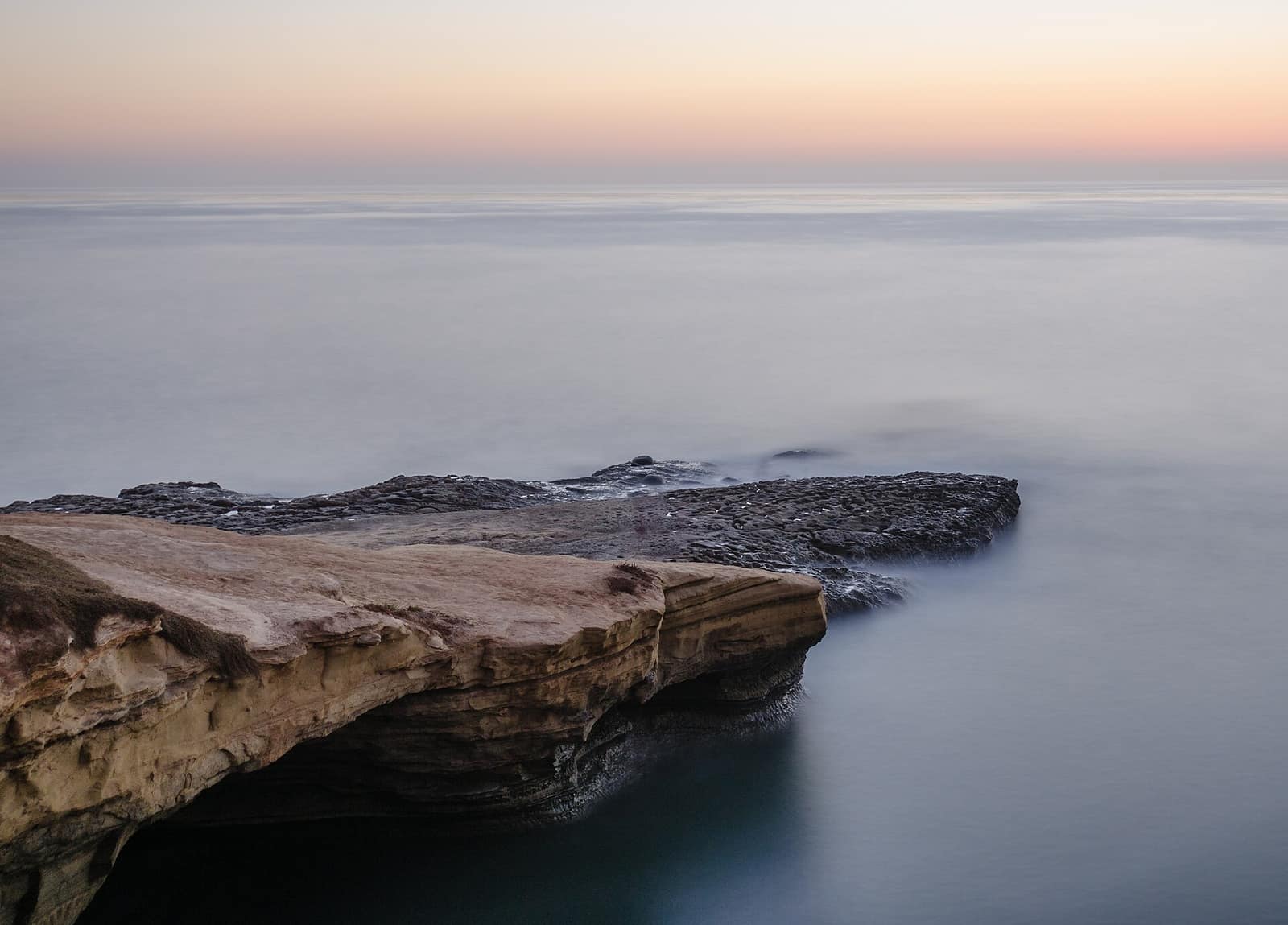 Layered sandstone cliffs at Sunset Cliffs Natural Park in San Diego at dusk, with calm Pacific Ocean and pastel sky